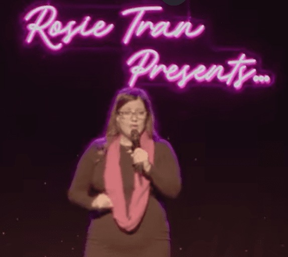 A woman performs stand-up comedy with neon sign 'Rosie Tran Presents' behind her.