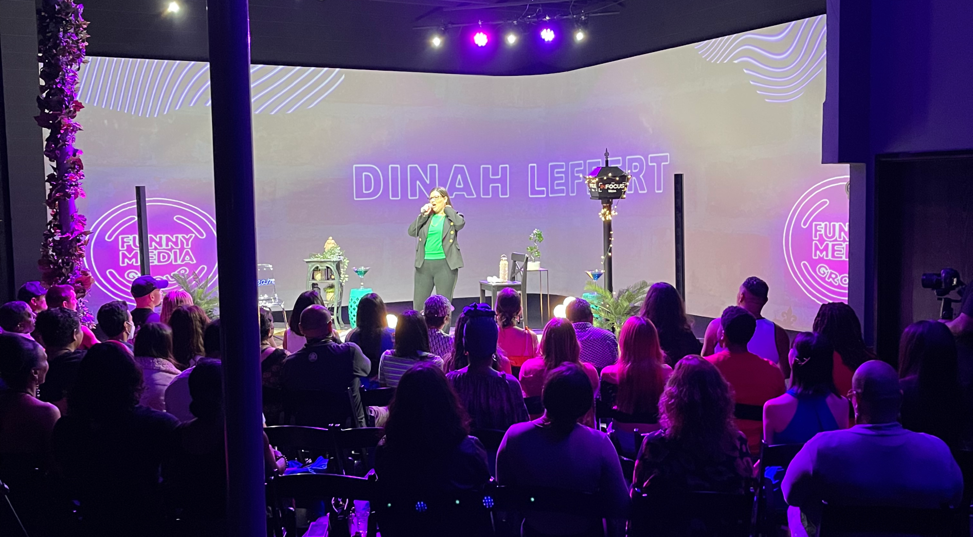 Dimah Lee performs on stage to a seated audience in a vibrant purple-lit venue.